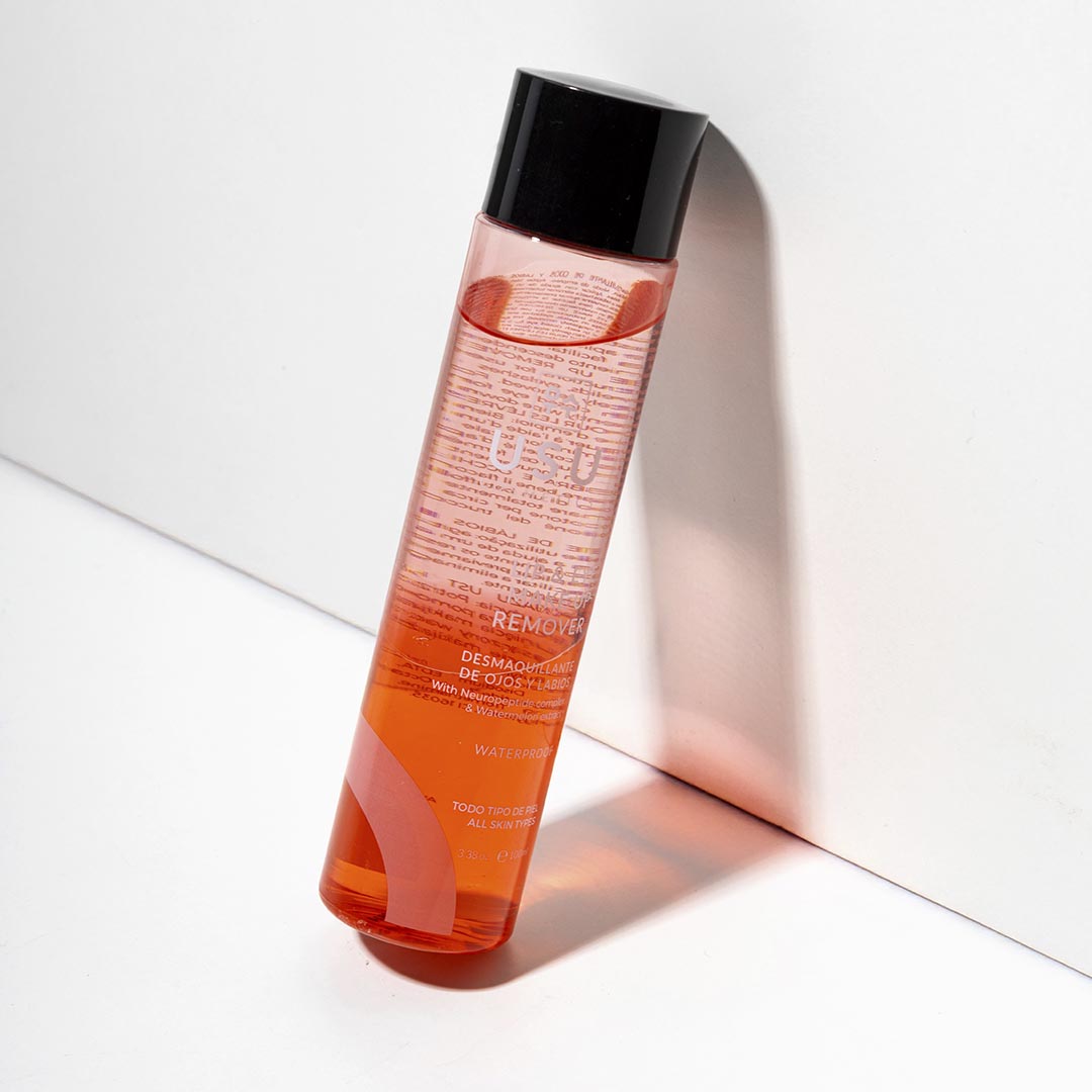 Botella de desmaquillante de ojos y labios apoyada en pared sobre fondo blanco. 